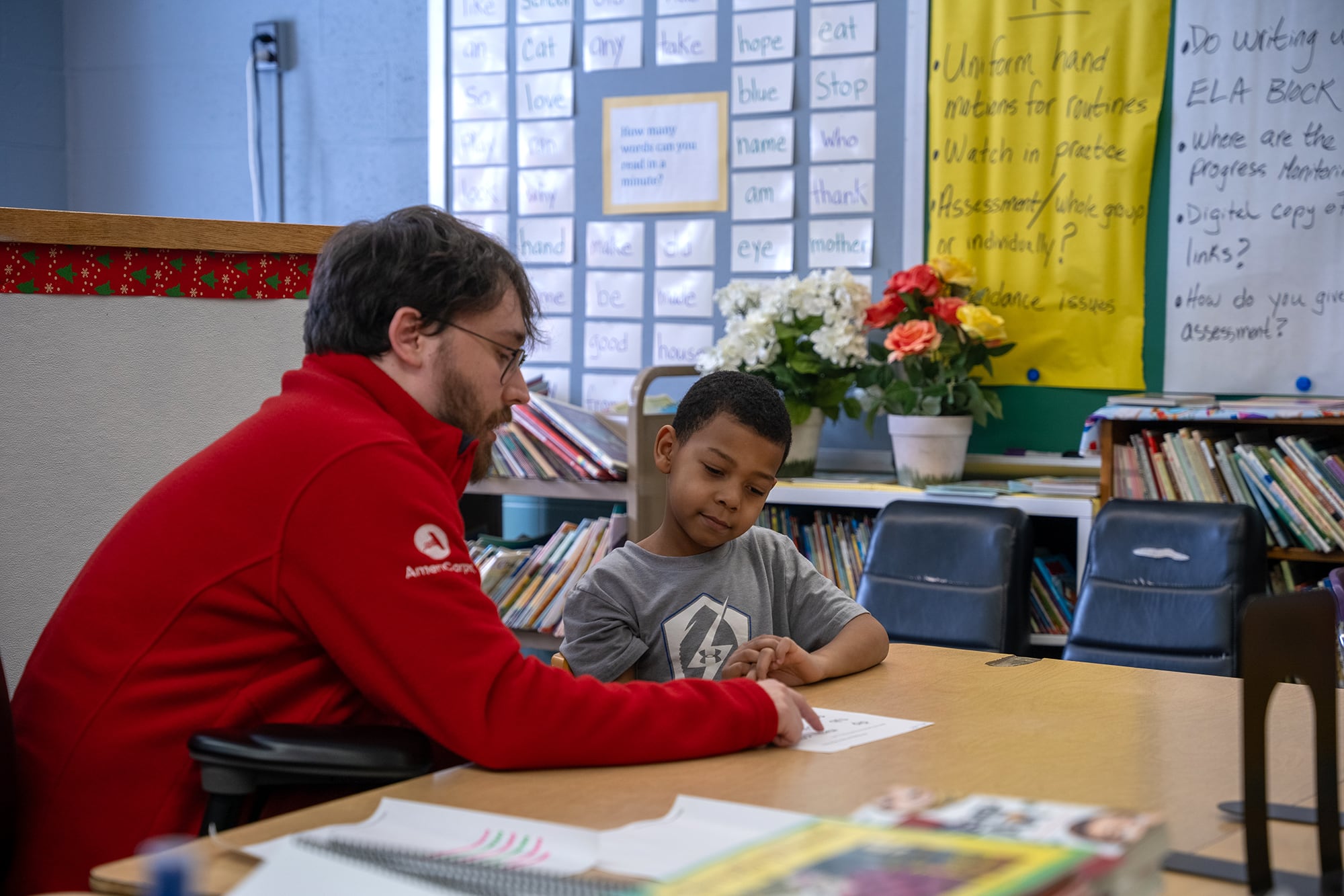 A photograph of a white man in a red sweater sitting next to a young Black student working at a desk in a classroom.