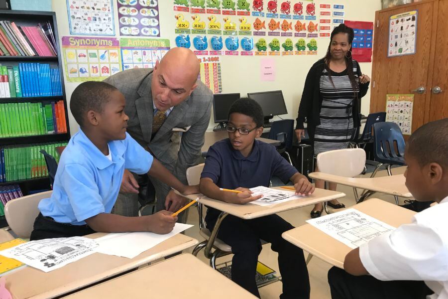 A man in a gray suit bends down to talk to a boy in a blue shirt as another boy in a blue shirt and a woman in gray and black look on.