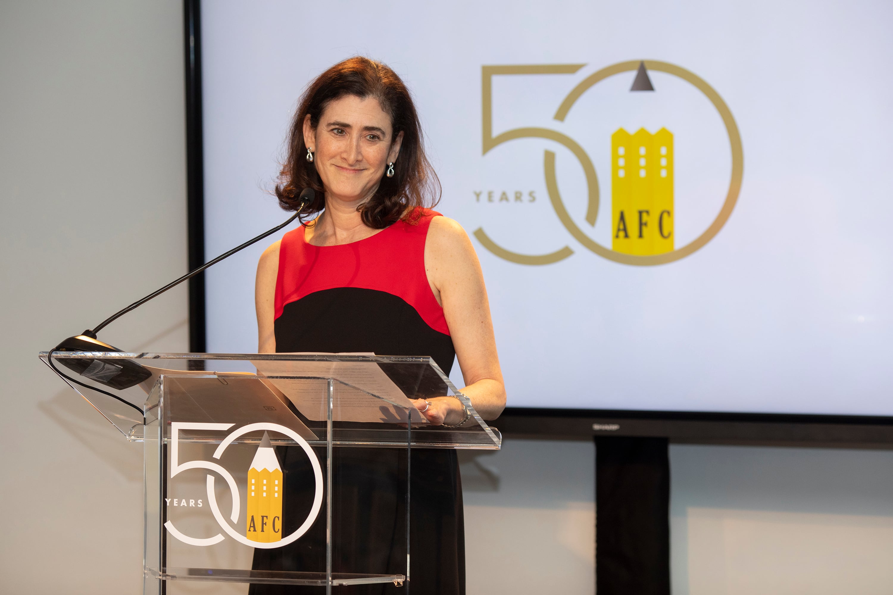 A photograph of a white woman in a red and black dress speaking from behind a podium and in front of a white screen with a gold and yellow logo.