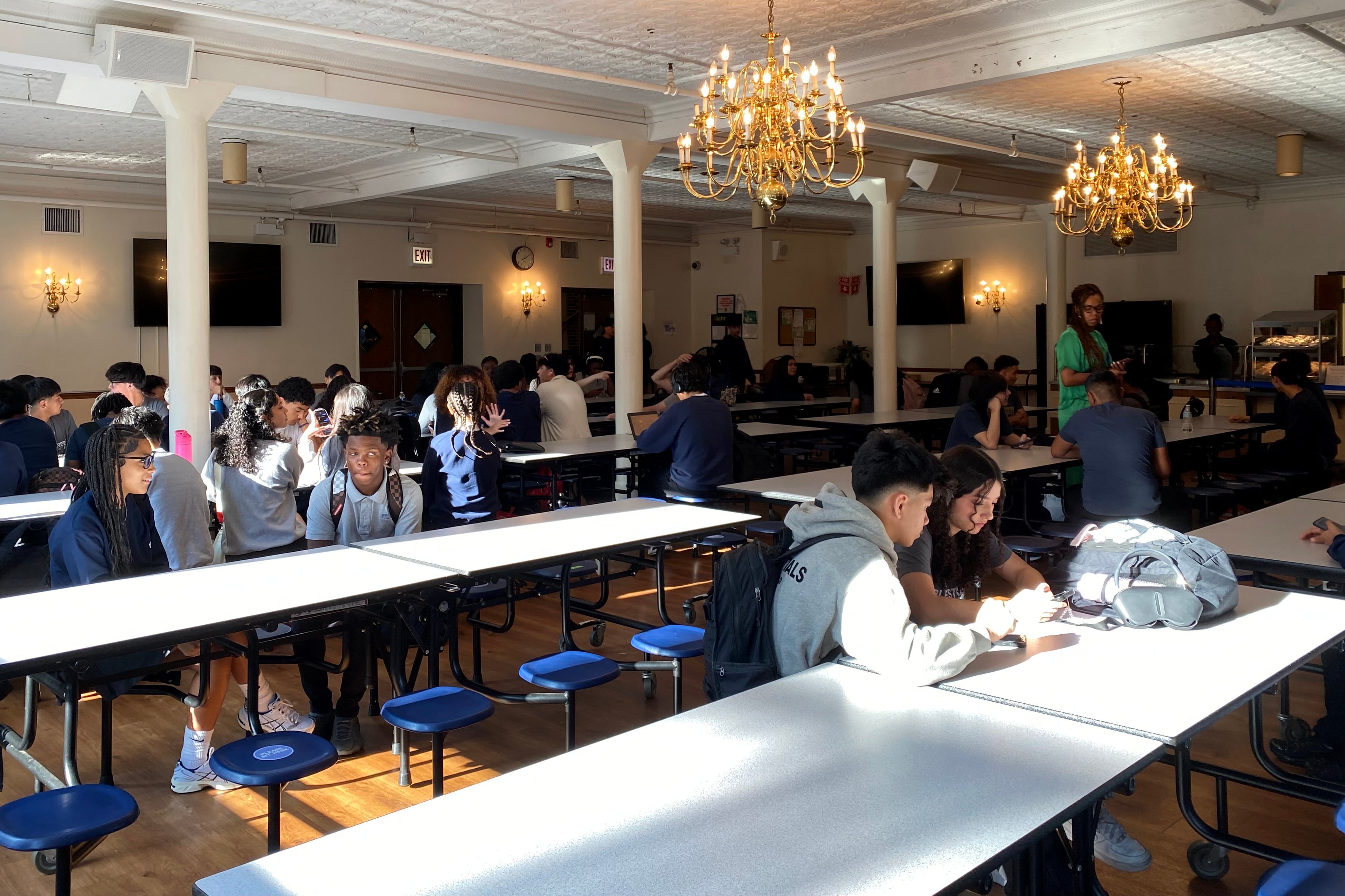 Students sit at tables in rows in a school cafeteria.