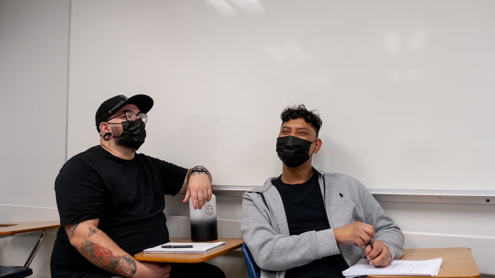 Two male students share a laugh together while sitting at their desks next to a white board. The student on the left is wearing a black hat and shirt, while the student on the right is wearing a grey hooded sweatshirt. They are both wearing black protective masks.