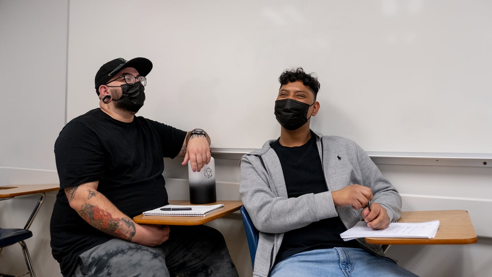 Two male students share a laugh together while sitting at their desks next to a white board. The student on the left is wearing a black hat and shirt, while the student on the right is wearing a grey hooded sweatshirt. They are both wearing black protective masks.