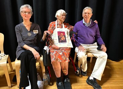 A woman in a flowered dress sits between a woman with a gray top and black pants and a man in a purple msweat and light pants