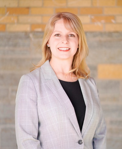 A photograph of a white woman in a suit jacket with medium length blonde hair posing for a photograph and smiling.
