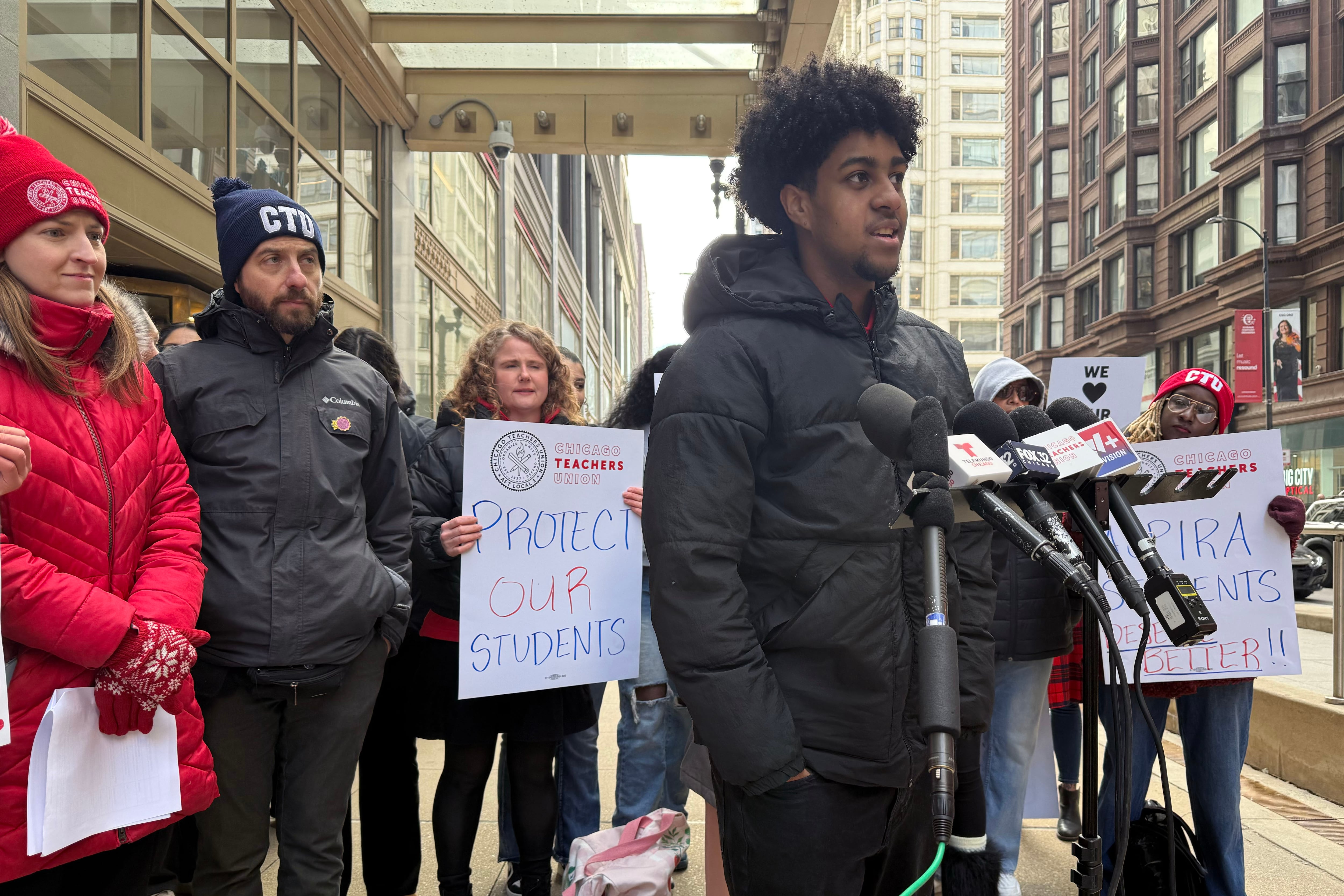A photograph of a Black high school student wearing a winter coat speaks into a microphone outside with a large group of people standing behind him.