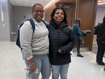 An adult and a young student pose for a portrait in a hallway.