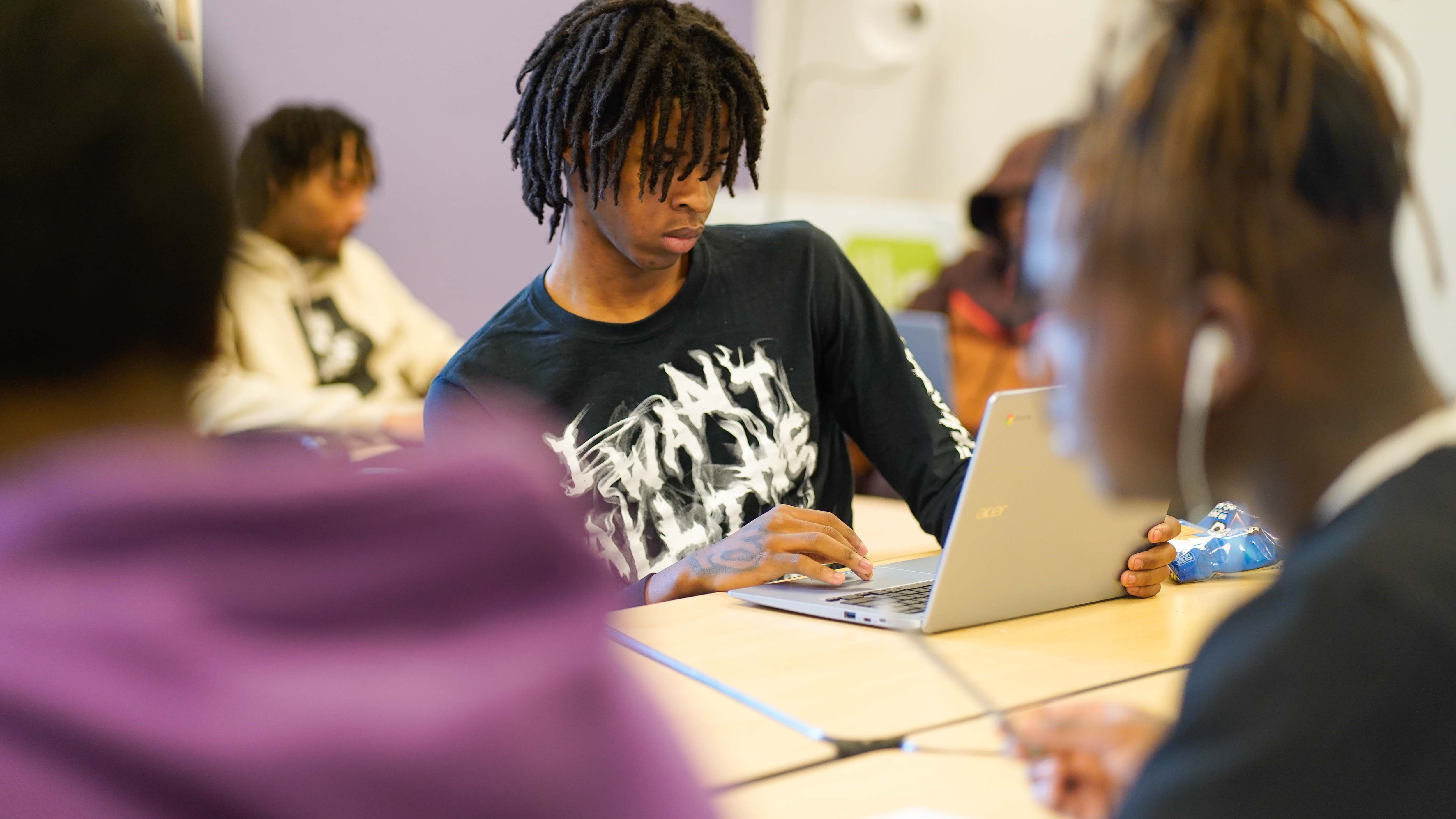 A teenage boy in a black t-shirt works at his computer surrounded by other students.