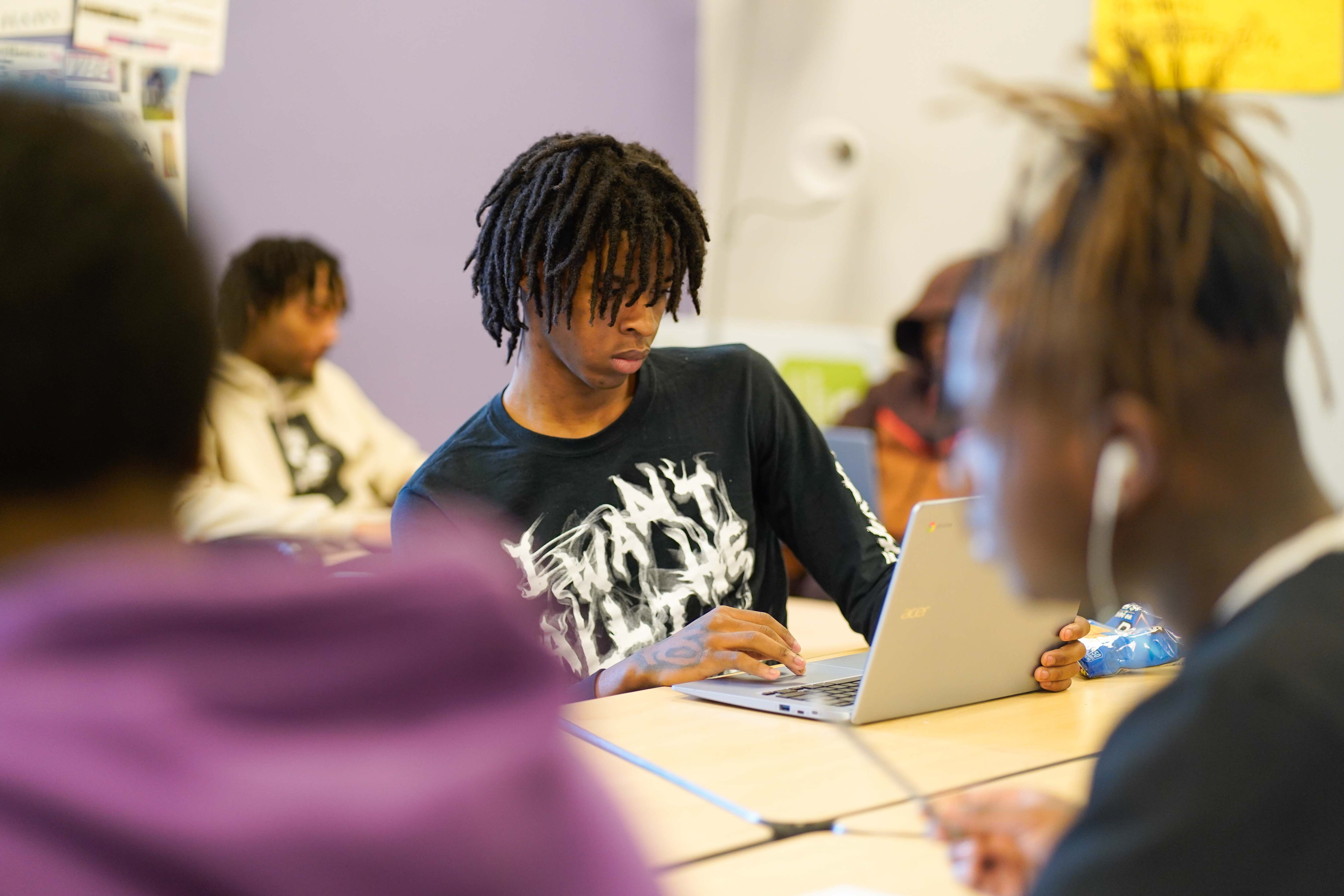 A teenage boy in a black t-shirt works at his computer surrounded by other students.