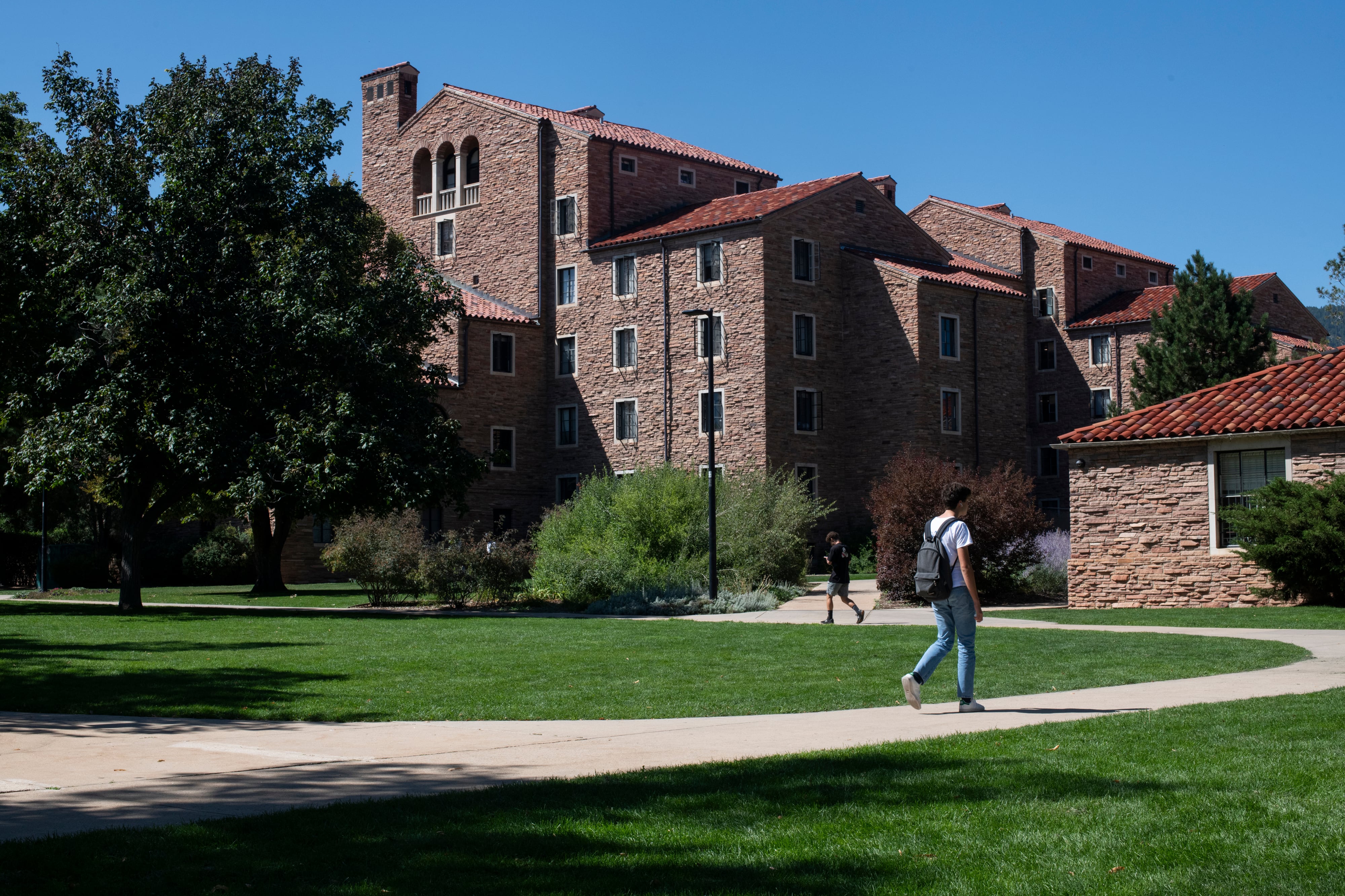 Two students walk on a concrete path in front of a large university campus with green grass and green trees all around.