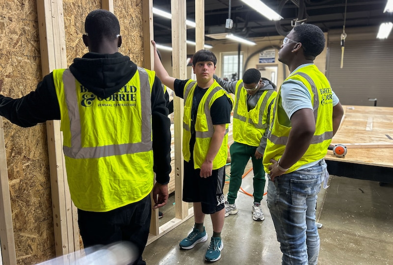 Four young men in brightly-colored bests stand in a room that looks like a woodshop.