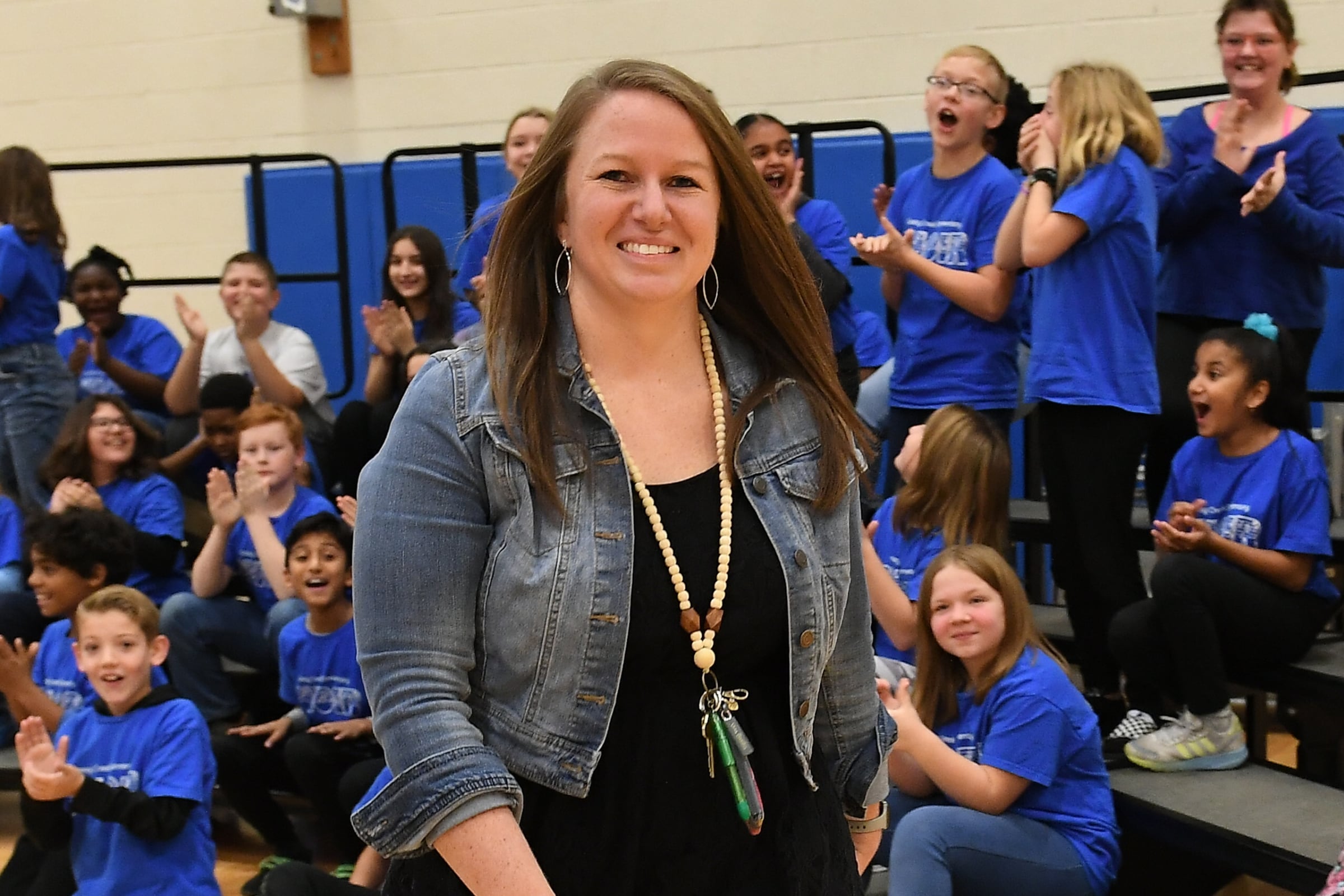Angela Fowler is shown in a denim jacket and black top as she walks past bleachers full of students in blue t-shirts to accept an award.