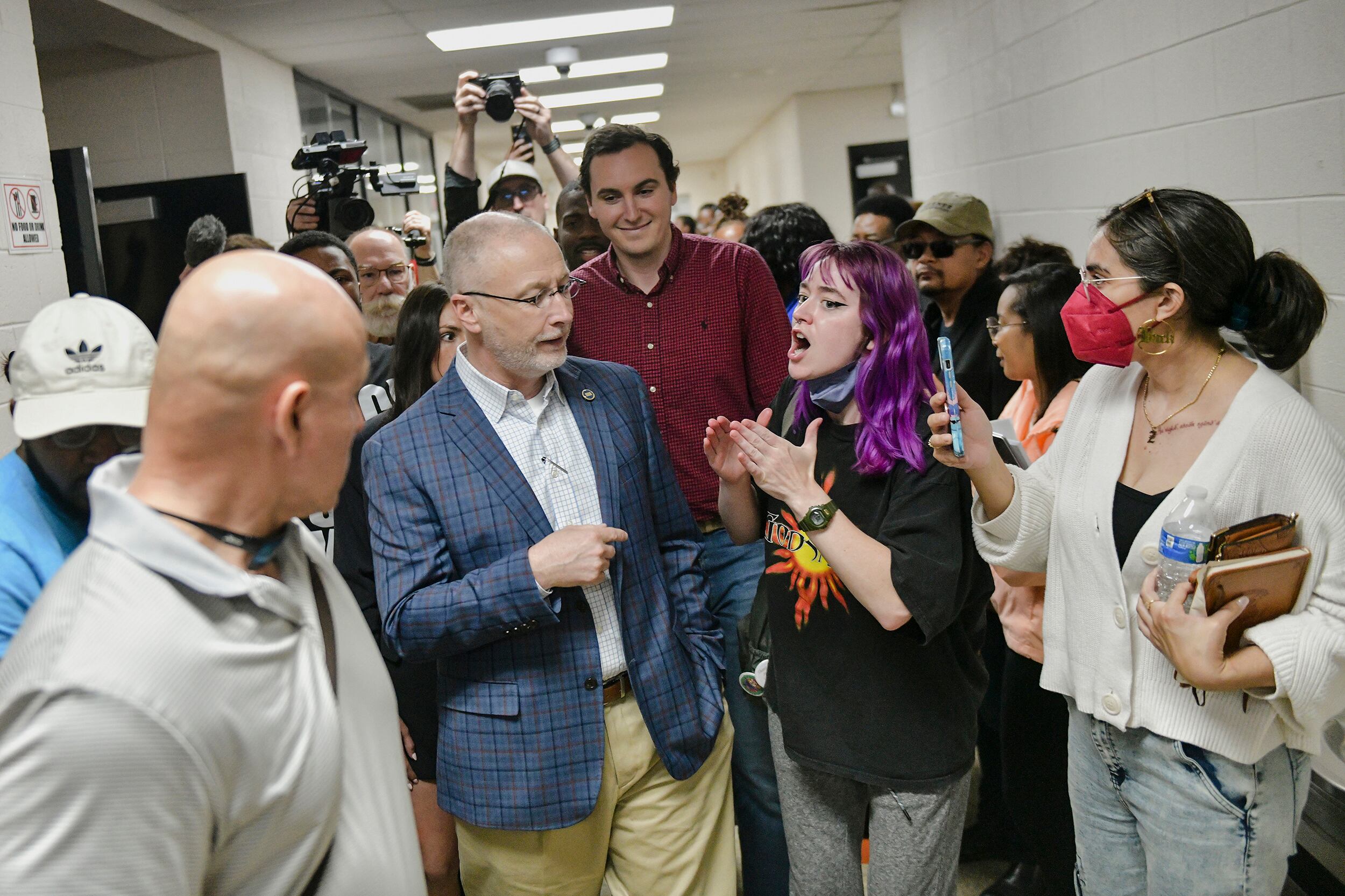 A photograph of a large group of people in a hallway. There's a young person with purple hair talking with an older white man with a suit jacket while a group of photojournalists in the back trying to make photos.
