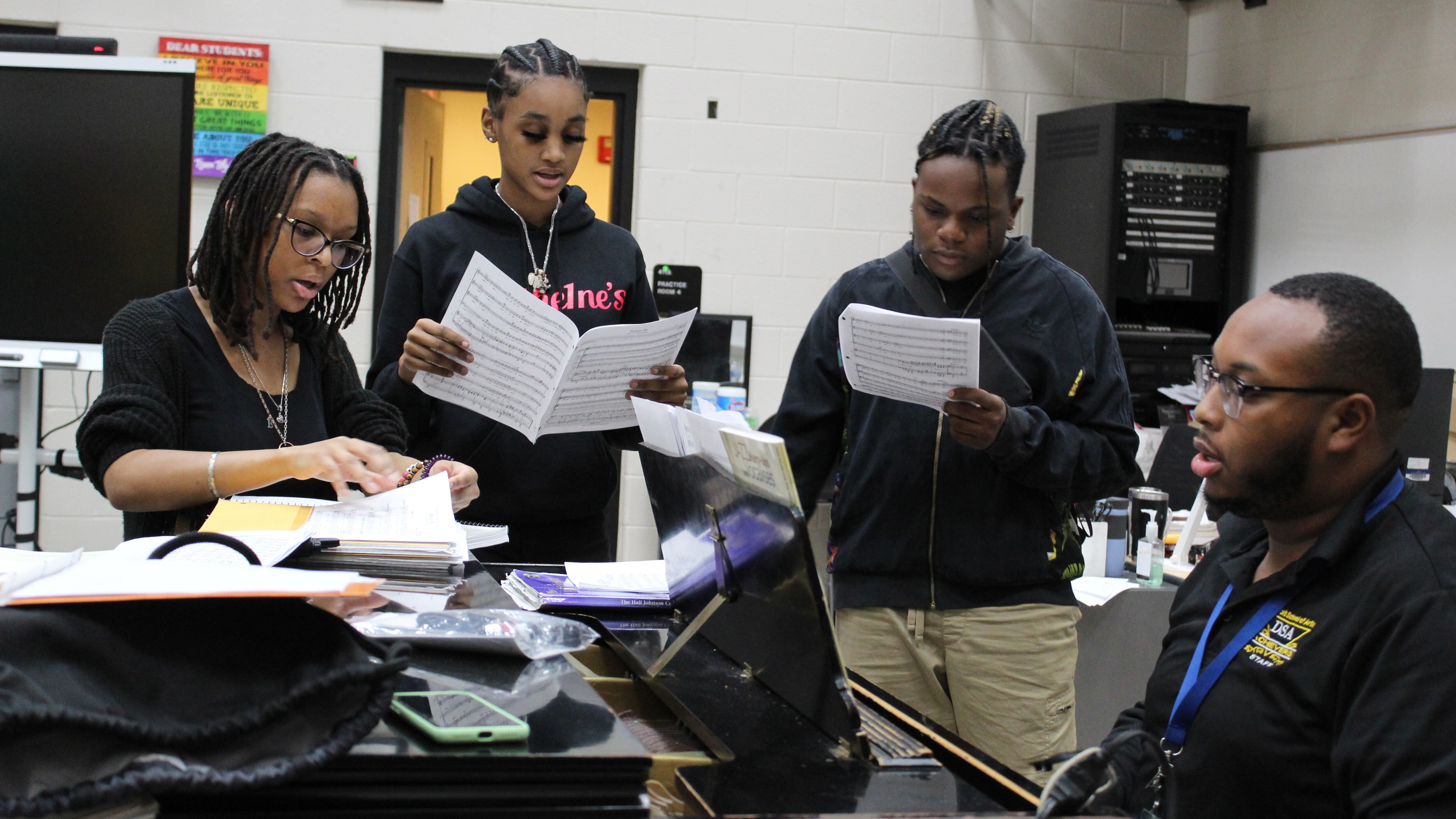 Three students stand on the side of a piano, singing while holding music sheets as their teacher plays the piano.