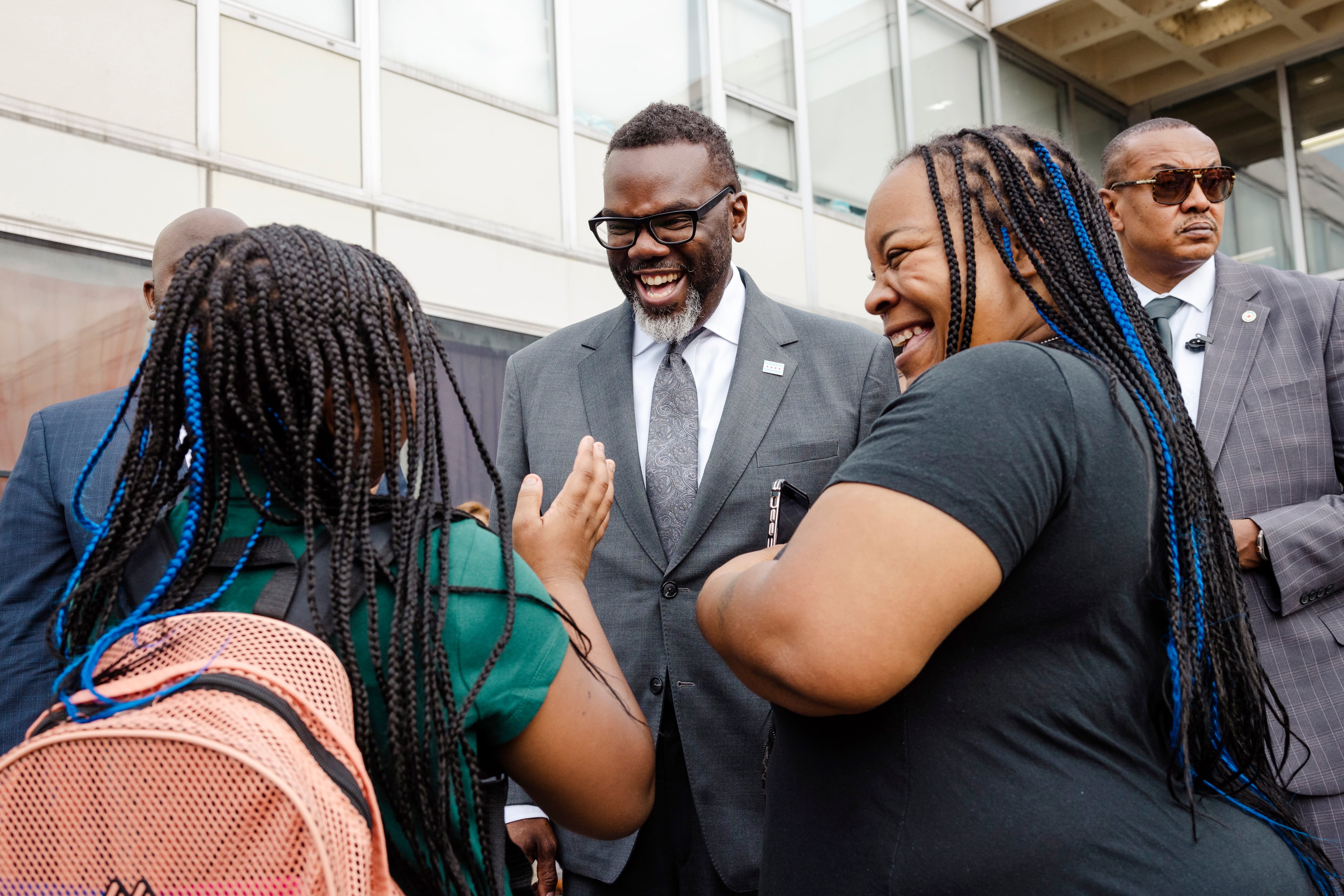 An adult in a suit talks with a young child and another adult outside of a school building on the first day of school.
