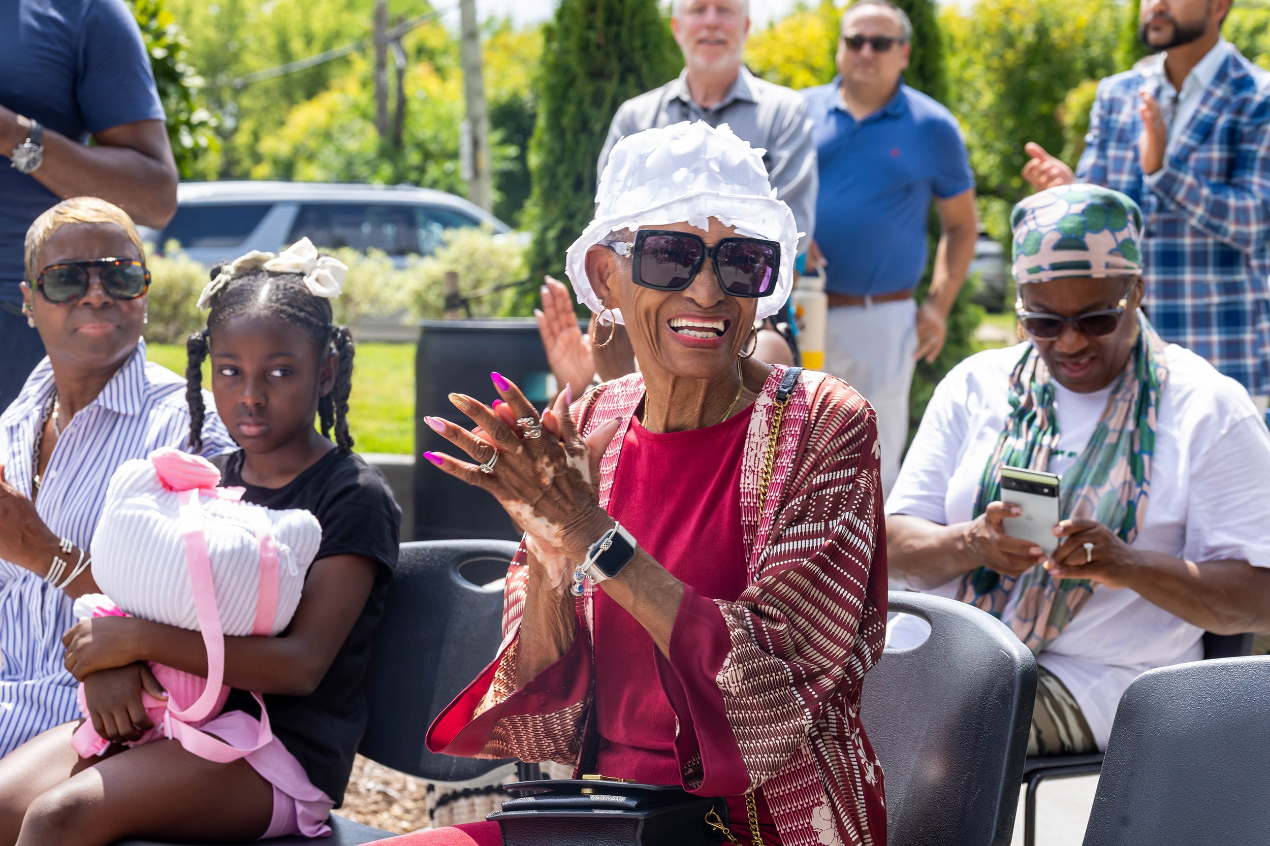 A photograph of a Black woman clapping while sitting at a chair in a crowd outside.
