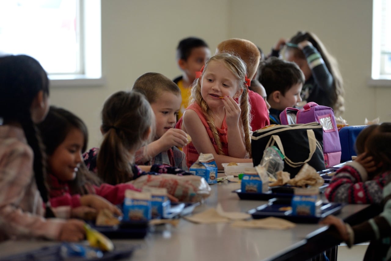Young students sit at a cafeteria table eating and talking. A blond girl turns to talk to her neighbor, a boy with short cropped hair.