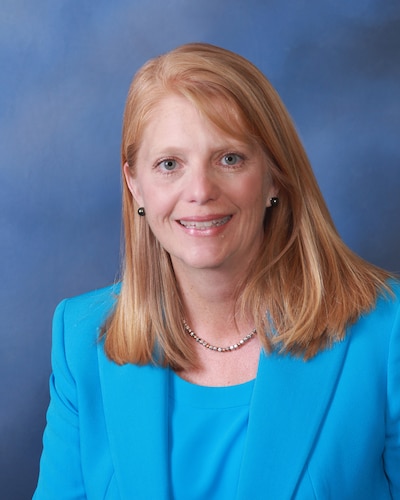 A woman with medium length orange hair and wearing a blue suit poses for a portrait in front of a blue background.