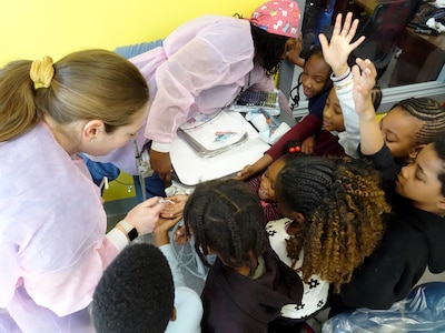 Two adults wearing scrubs show off dentist equipment to a large group of young students.