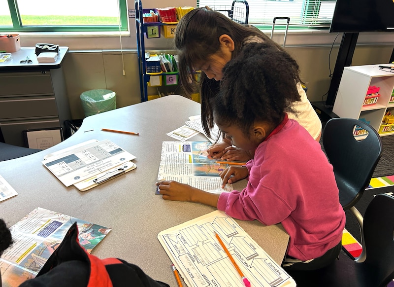 Two young students work at a table in front of a window.