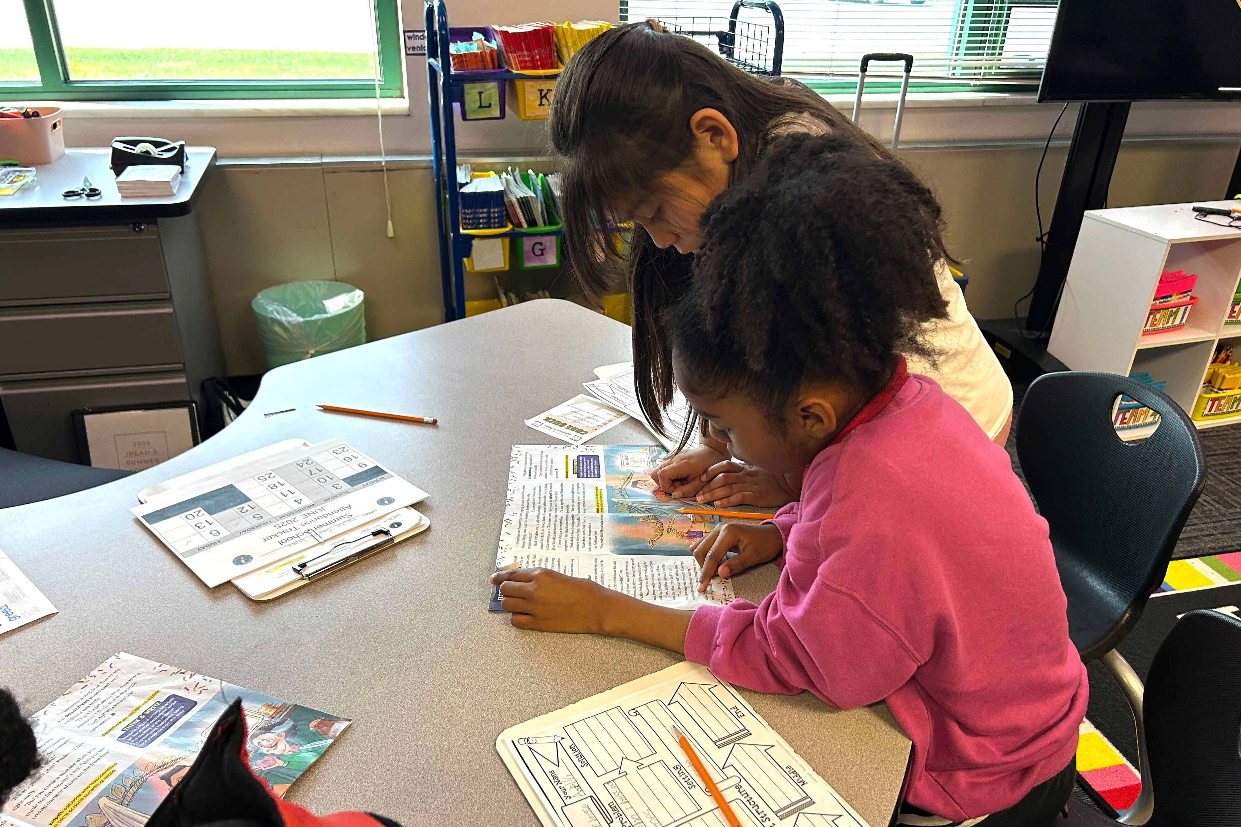 Two young students work at a table in front of a window.