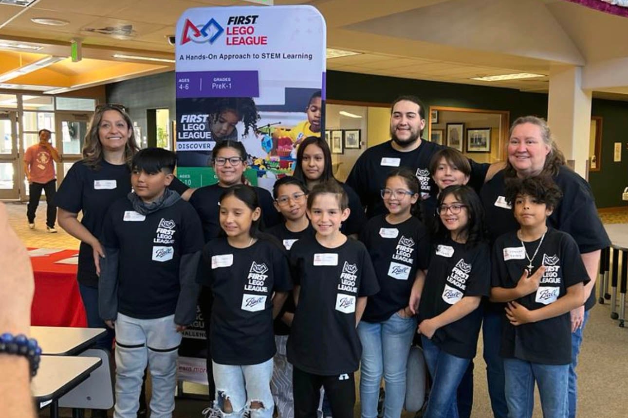 A group young students and three adults wearing matching black shirts and pose for a group photo with a banner and a school hallway in the background.