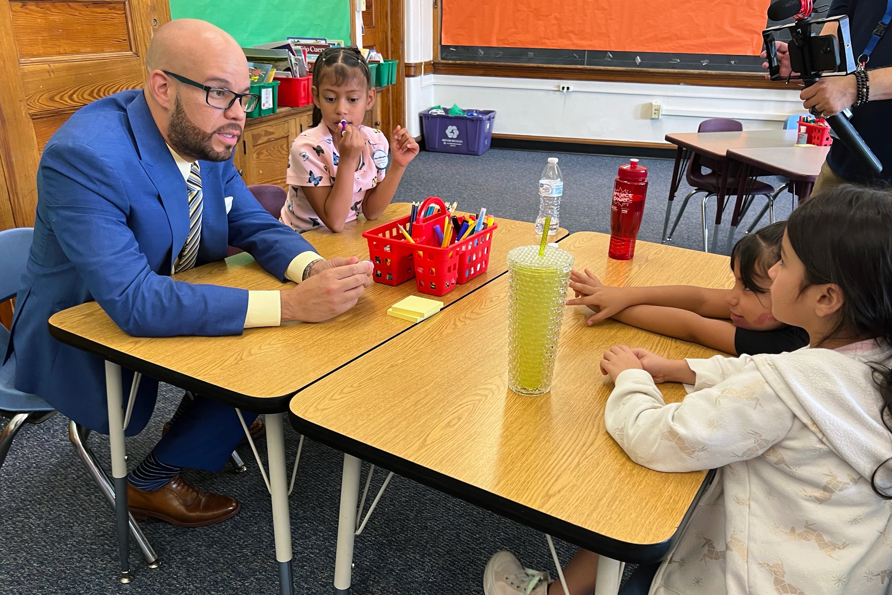 An adult in a blue suit sits next to three young students in a classroom.