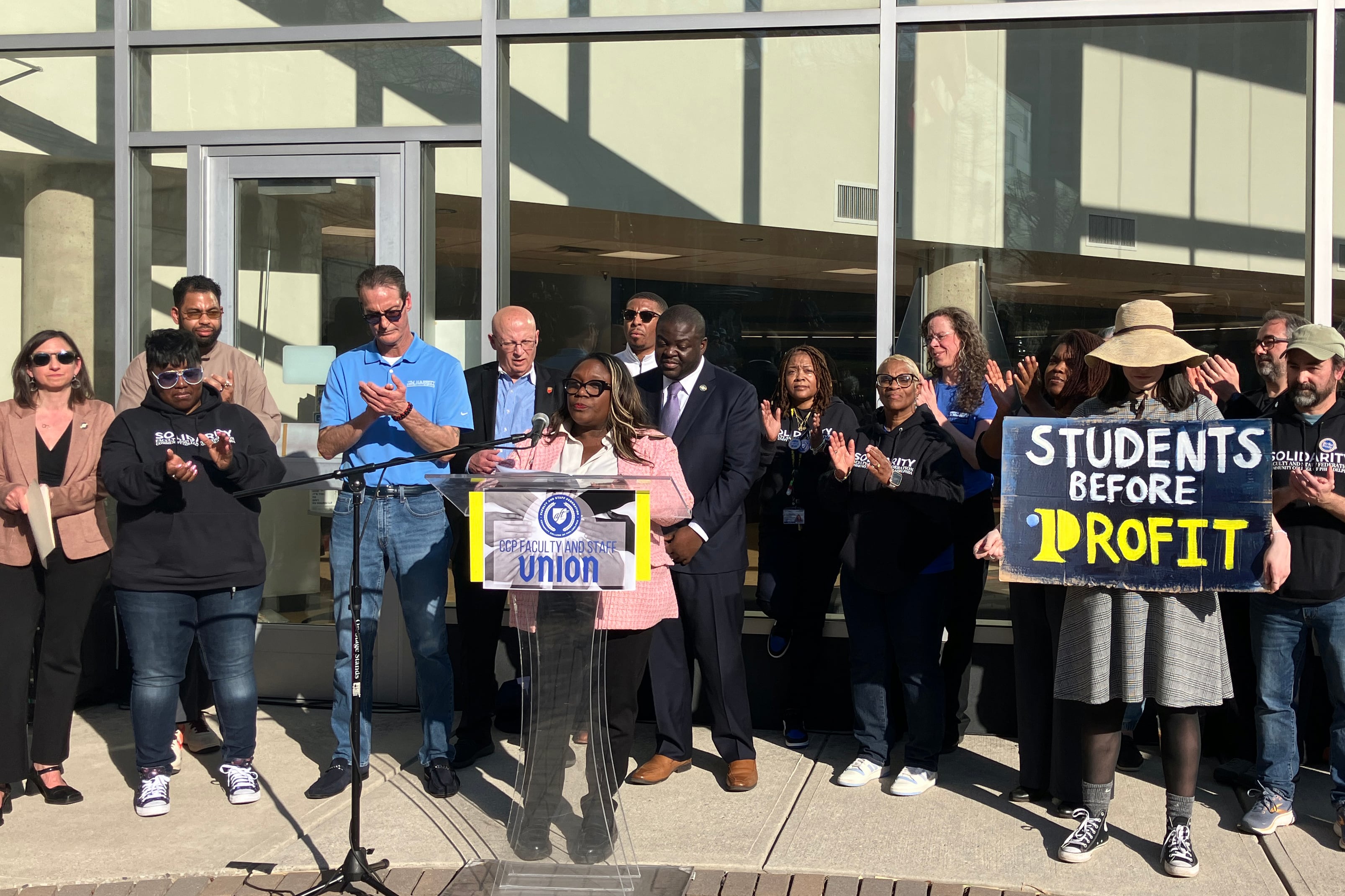 A person speaks from behind a small podium with a large group of people standing behind in support. One person is holding a sign that reads "students before profit."