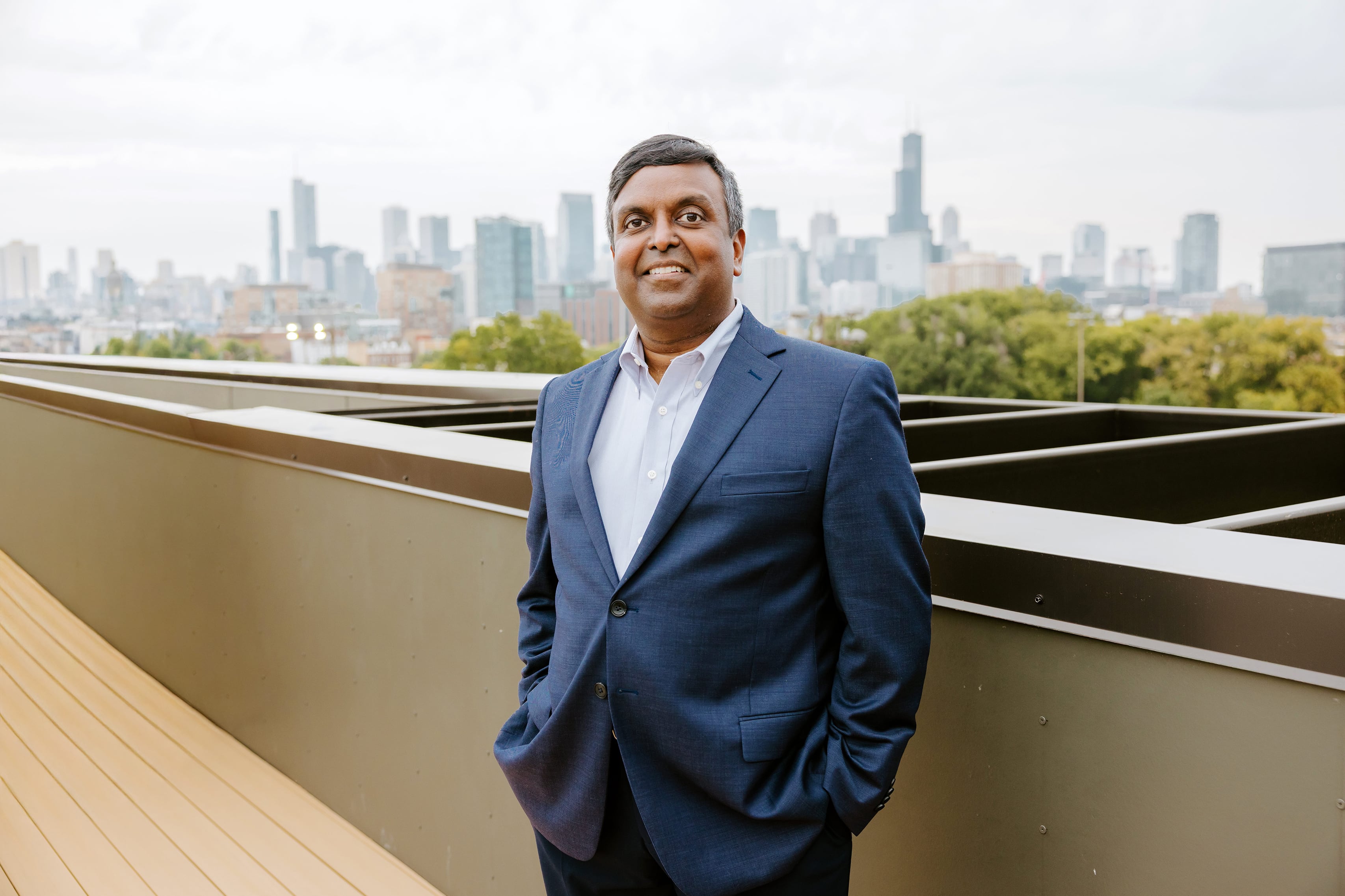 A photograph of a man in a blue suit smiling for a portrait with the city skyline in the background.