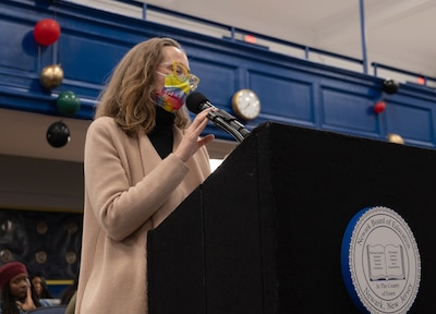 A photograph of a white woman wearing a tie dye face mask speaks from a podium in a school auditorium.