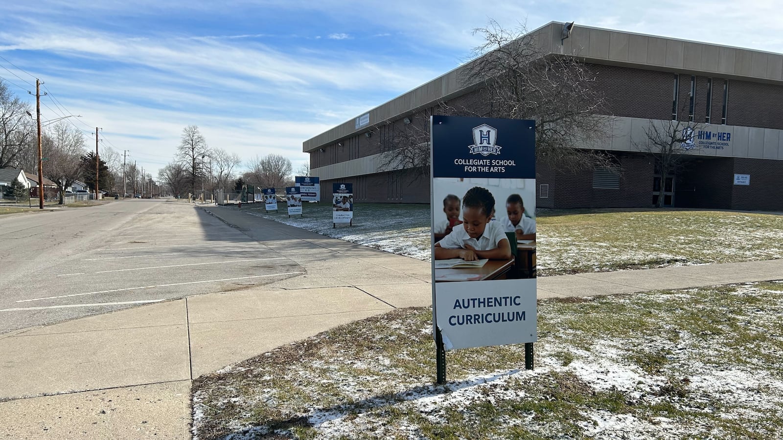 A sign reading “Him by Her Collegiate School for the Arts” stands in a snowy ground with similar signs in the background. In the background is a brick school building.