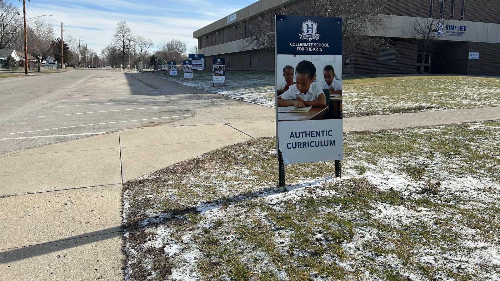 A sign reading “Him by Her Collegiate School for the Arts” stands in a snowy ground with similar signs in the background. In the background is a brick school building.
