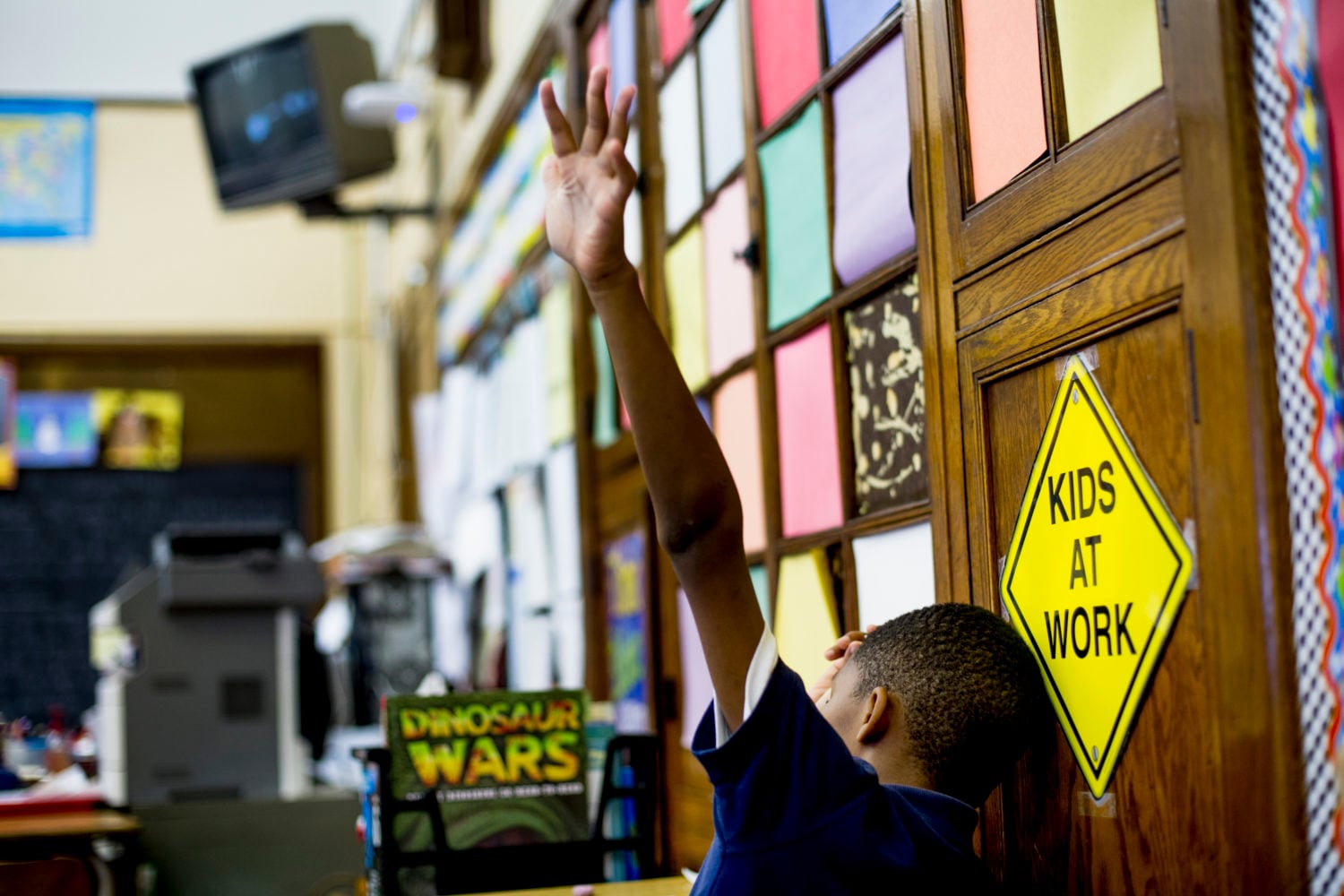 A student is shown holding their hand in the air while sitting at a desk in a classroom.