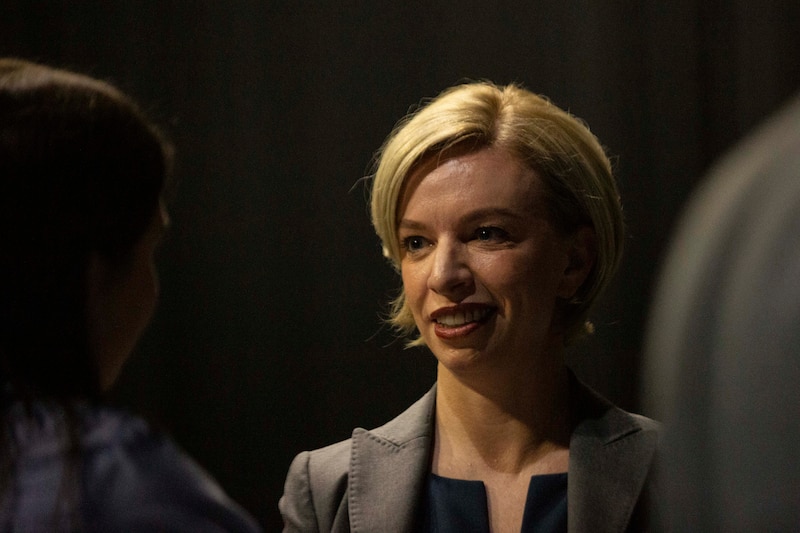 A white woman with short blonde hair speaks to a person in a dark room.