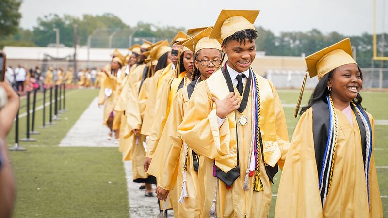 Students wearing gold graduation robes walk in a line for an outdoor ceremony.