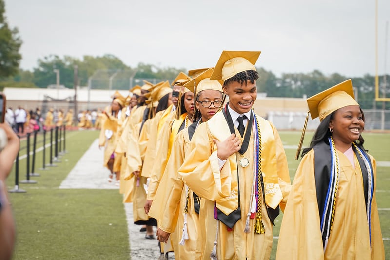 Students wearing gold graduation robes walk in a line for an outdoor ceremony.