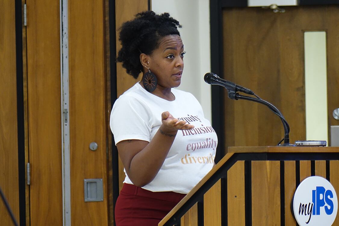 A woman with large, ornate earrings and a white shirt that reads “equity, inclusion, equality, diversity” speaks at a podium marked “myIPS”.