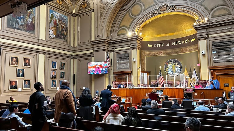 Several people stand in line while people sit in rows on each side of the line. A group of people sit at a wooden bench with the words "City of Newark" above them.