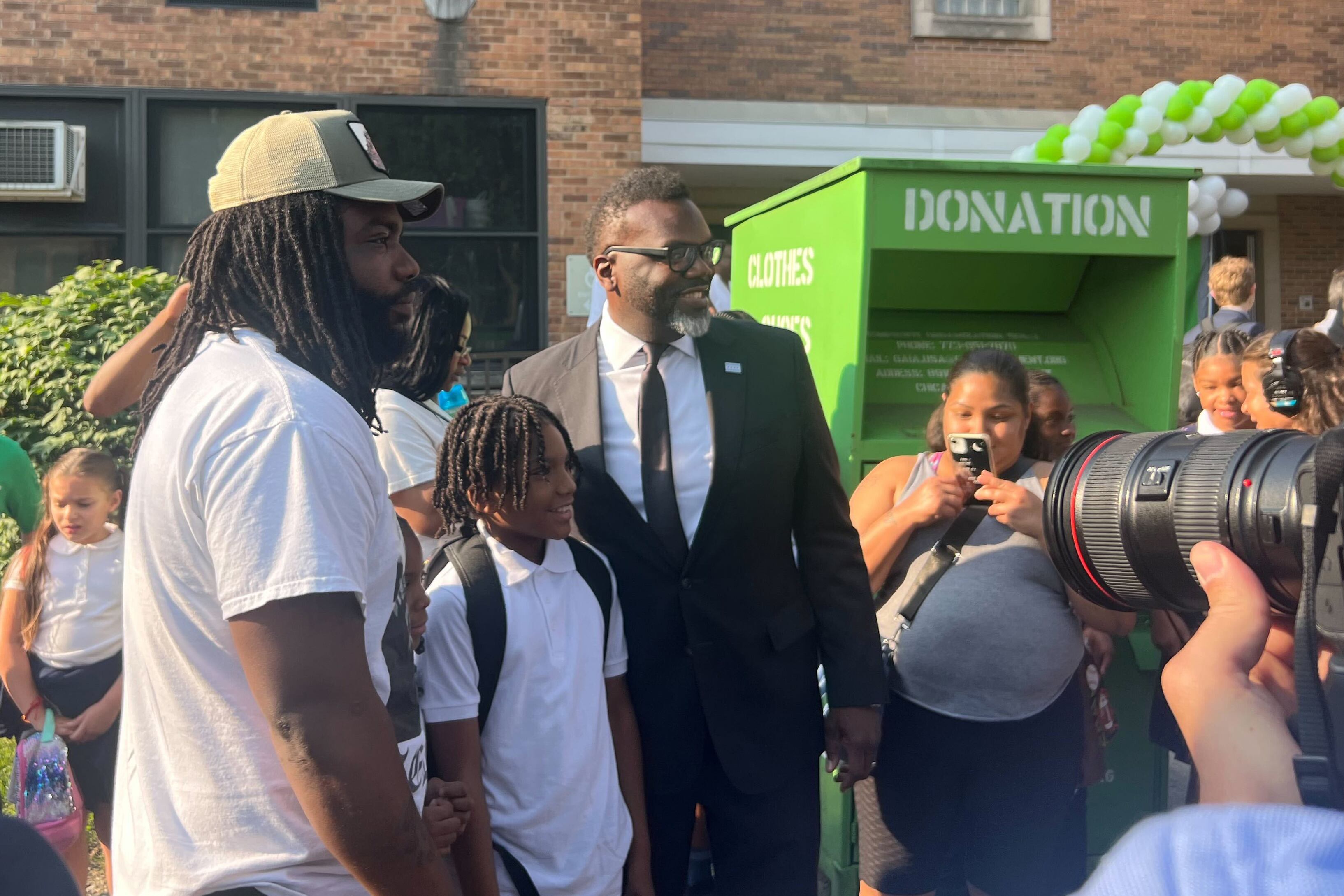 A man in a suit and tie greets elementary school children in front of cameras.