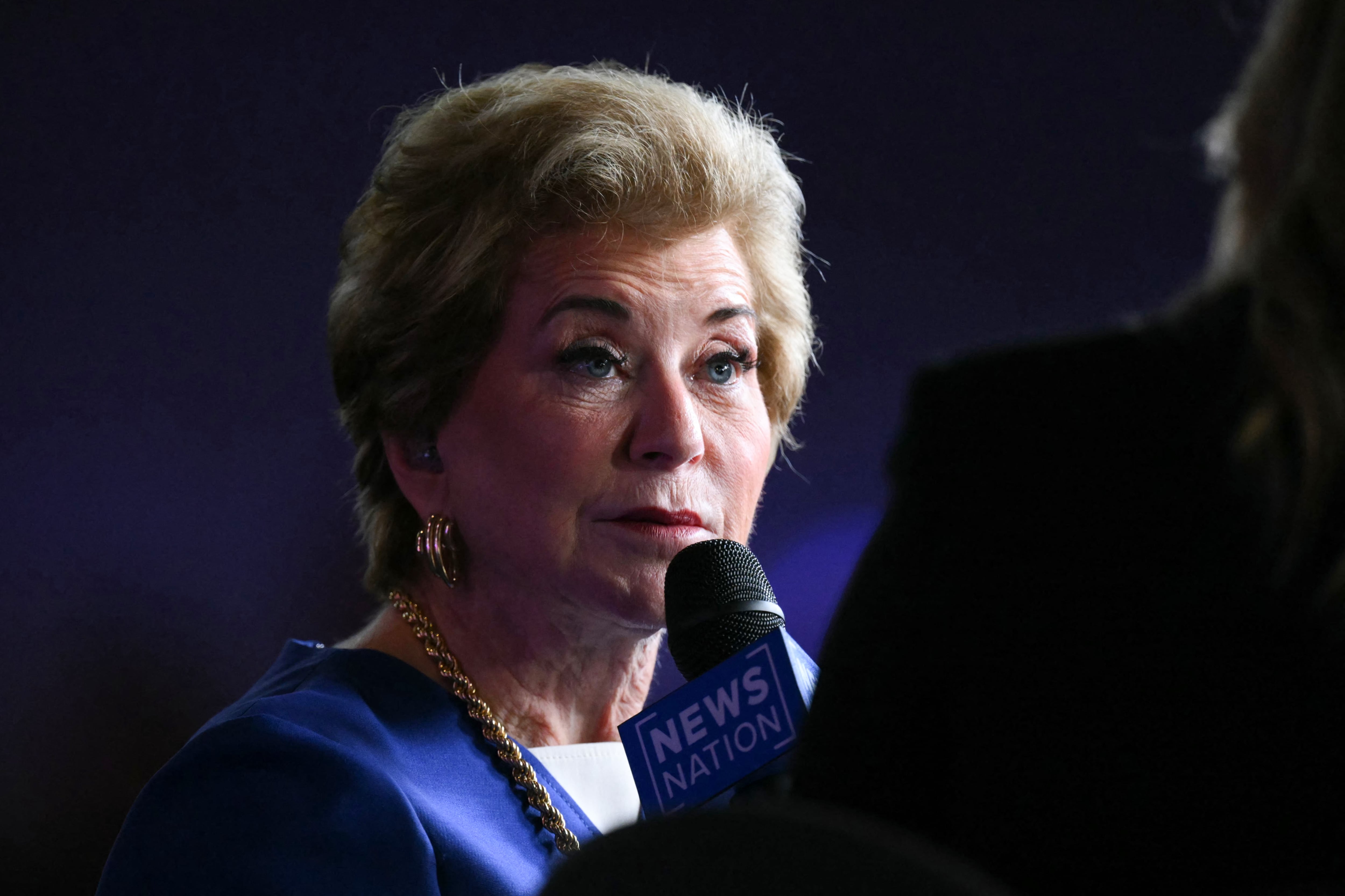A woman in a blue suit jacket speaks into a microphone.