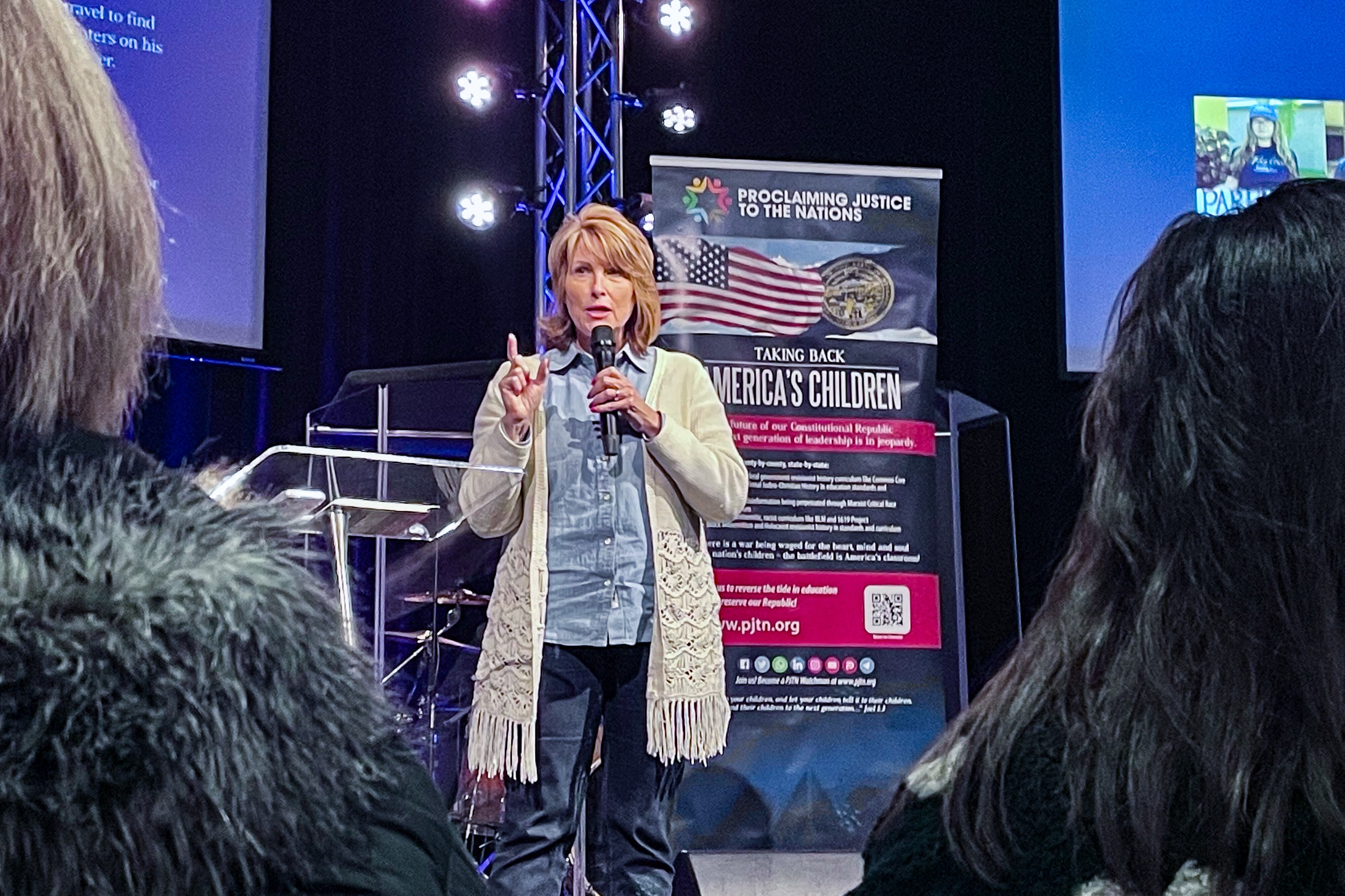 A woman speaks into a microphone on a stage in front of women at a meeting.