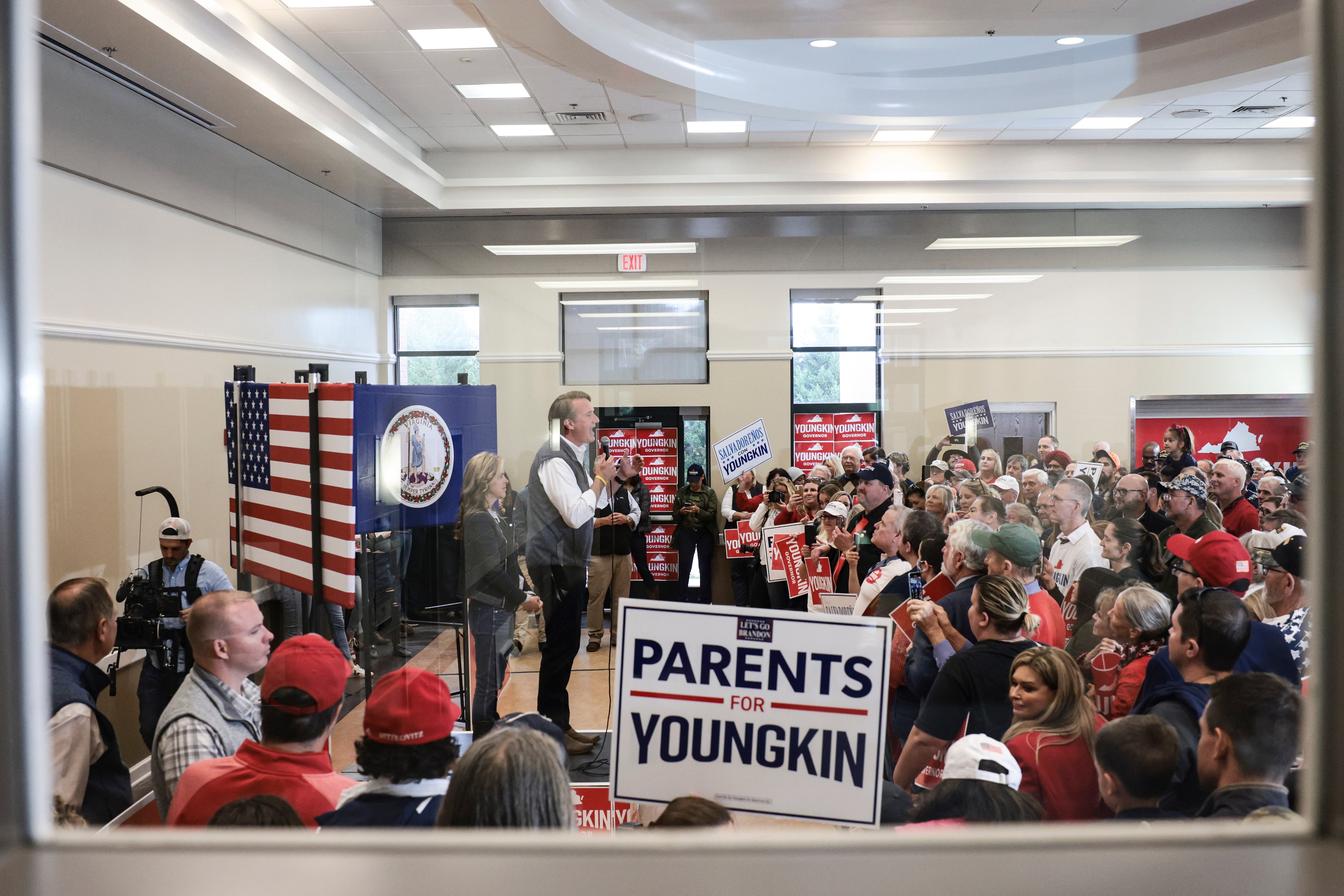 Virginia Governor-elect Glenn Youngkin speaks at an indoor rally, shown behind a glass window. A sign reading “Parents for Youngkin” sits in the foreground by the window.