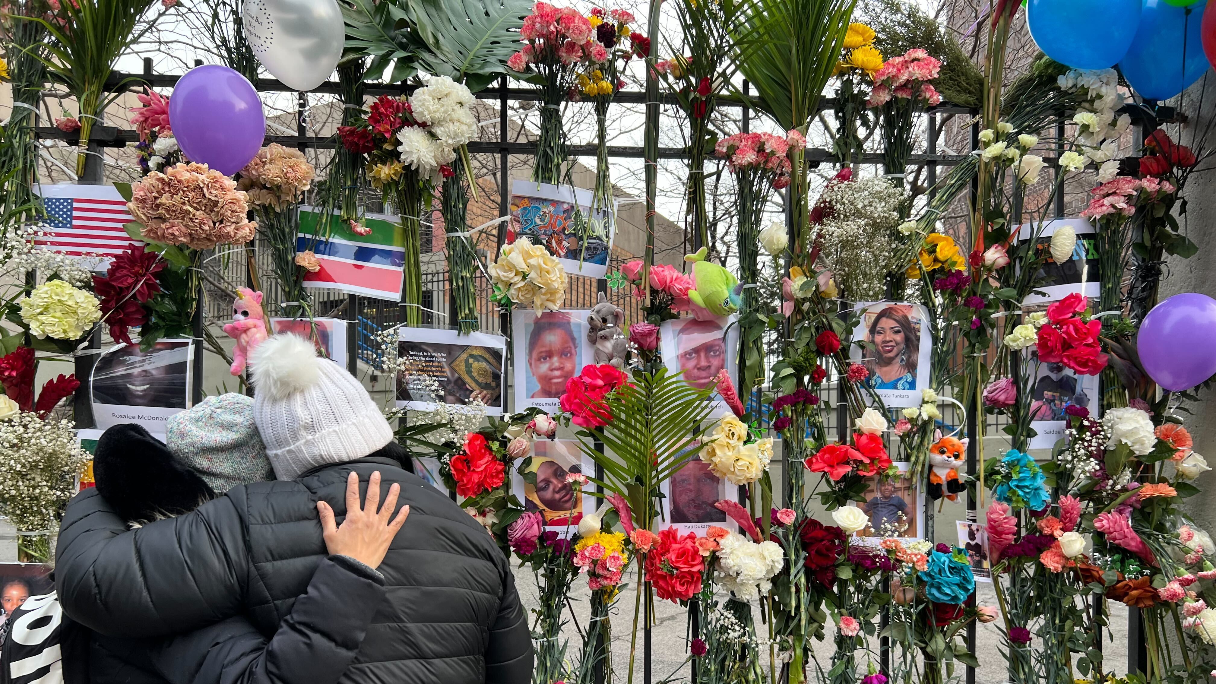 Two people in winter clothing embrace next to a memorial for the victims of a fatal fire in a Bronx apartment. The memorial is adorned with flowers, candles, and photographs of the victims.