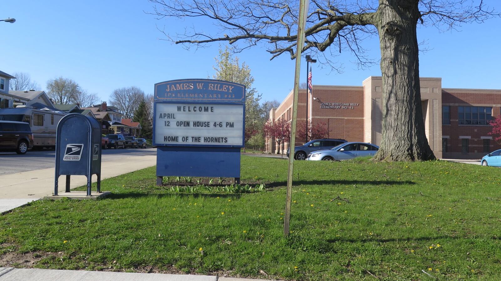 A blue school sign reading “James W. Riley” stands in a green lawn next to a blue postal box.