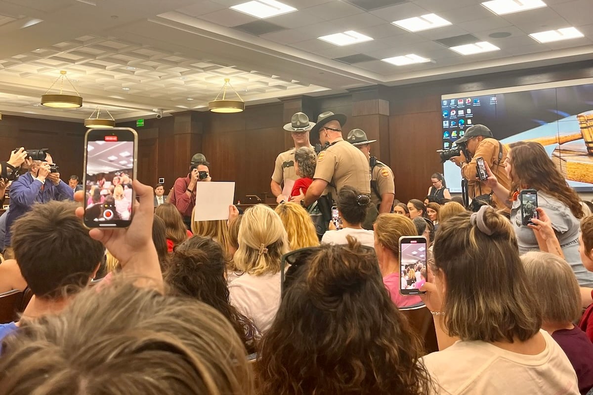 Three men wearing uniforms escort a woman from a crowded room.