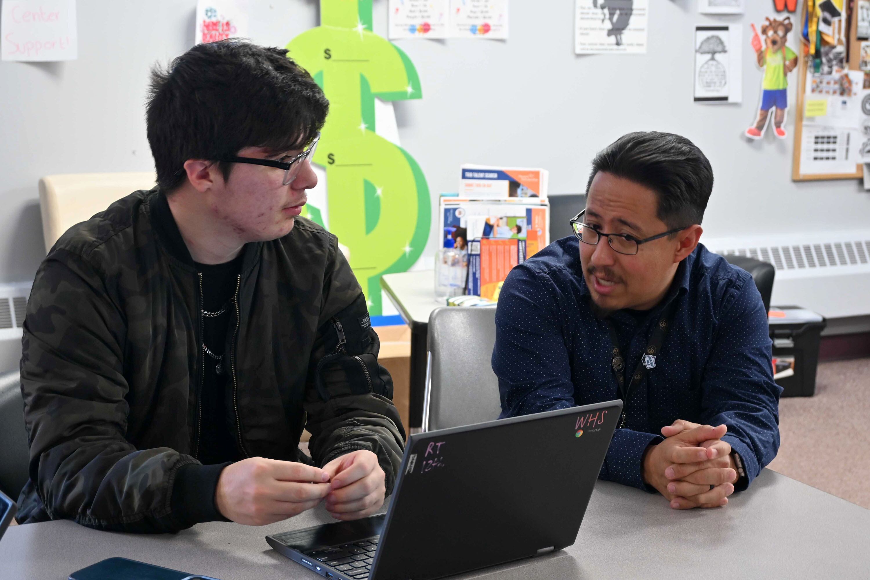 One high school student and one adult sit at a desk behind a laptop with a whiteboard in the background.