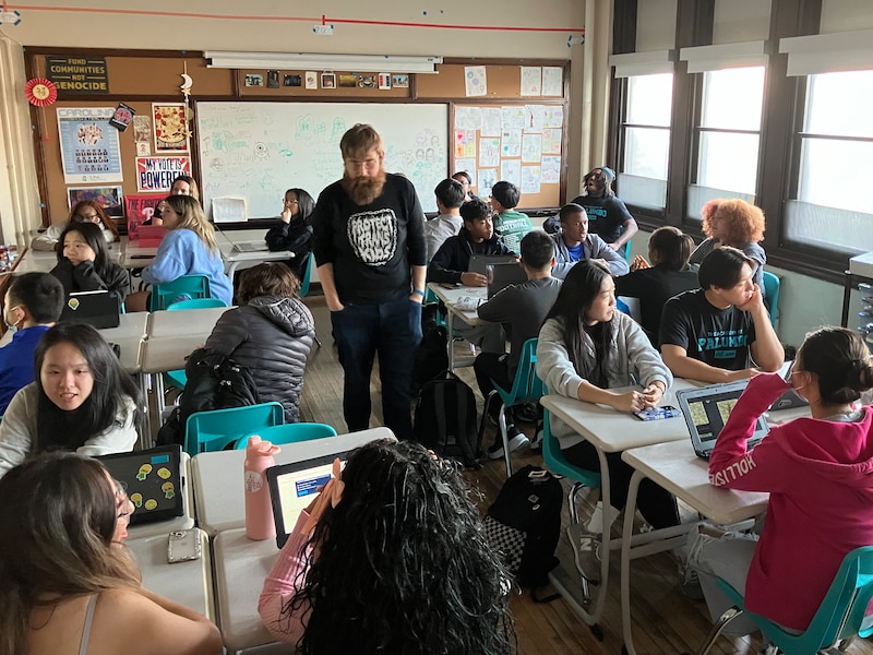 A teacher stands in the middle of a classroom filled with students at desks.