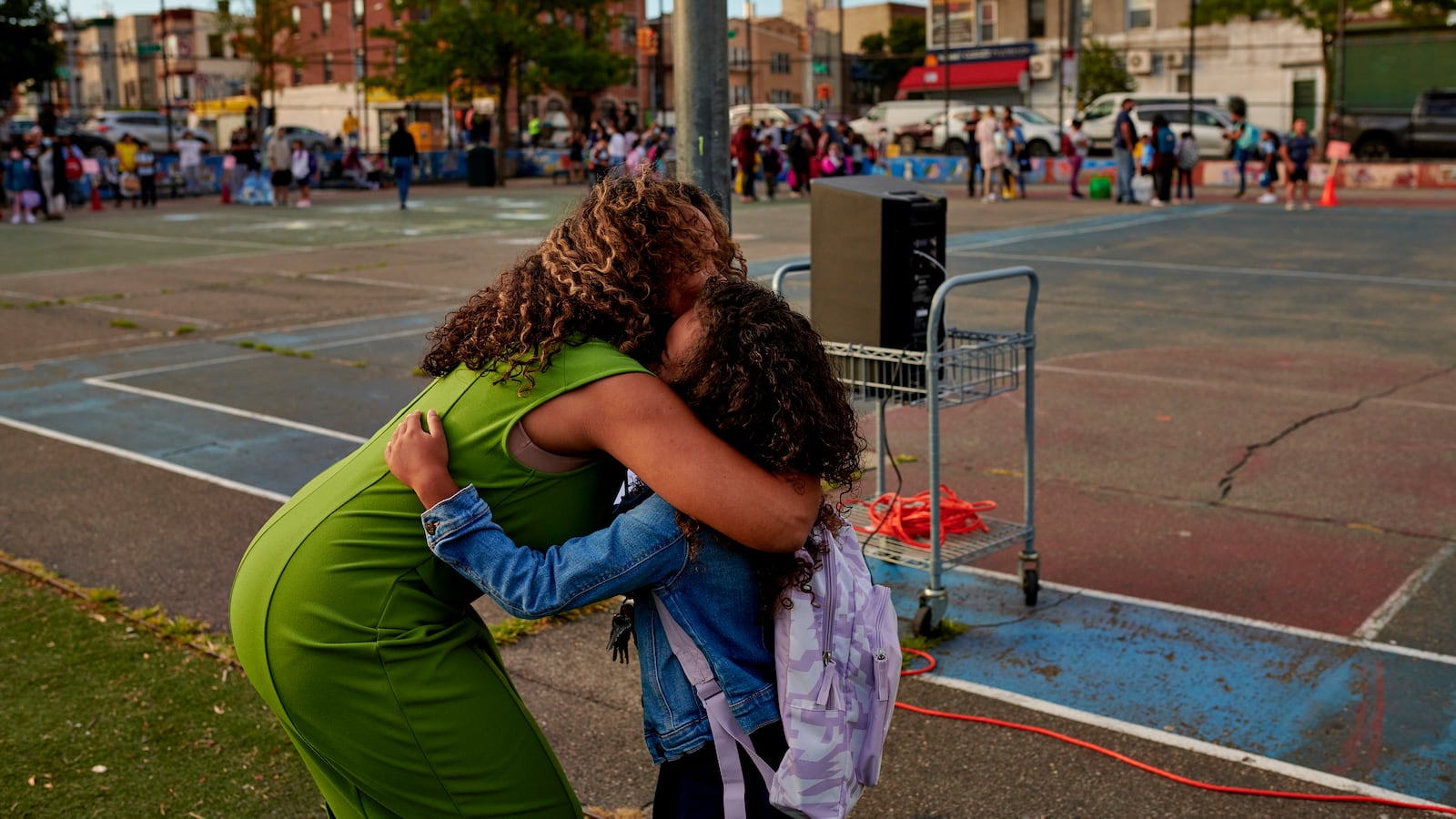 An adult woman wearing a bright green dress bends over to hug a student wearing a purple backpack and a jean jacket. Other students and an outdoor tennis court are in the background.