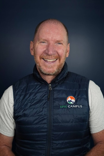 A photograph of a white man posing for a photograph in front of a dark background.