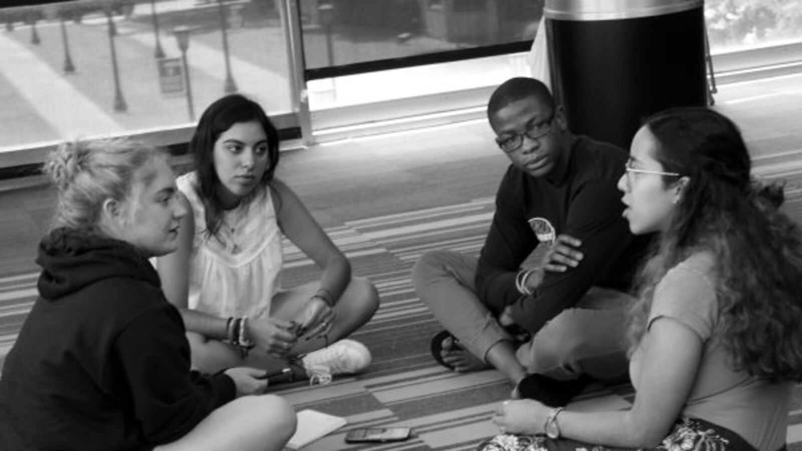 Four students sit on a classroom floor. One student is speaking and the other three are listening. They are participating in a Narrative 4 Story Exchange.