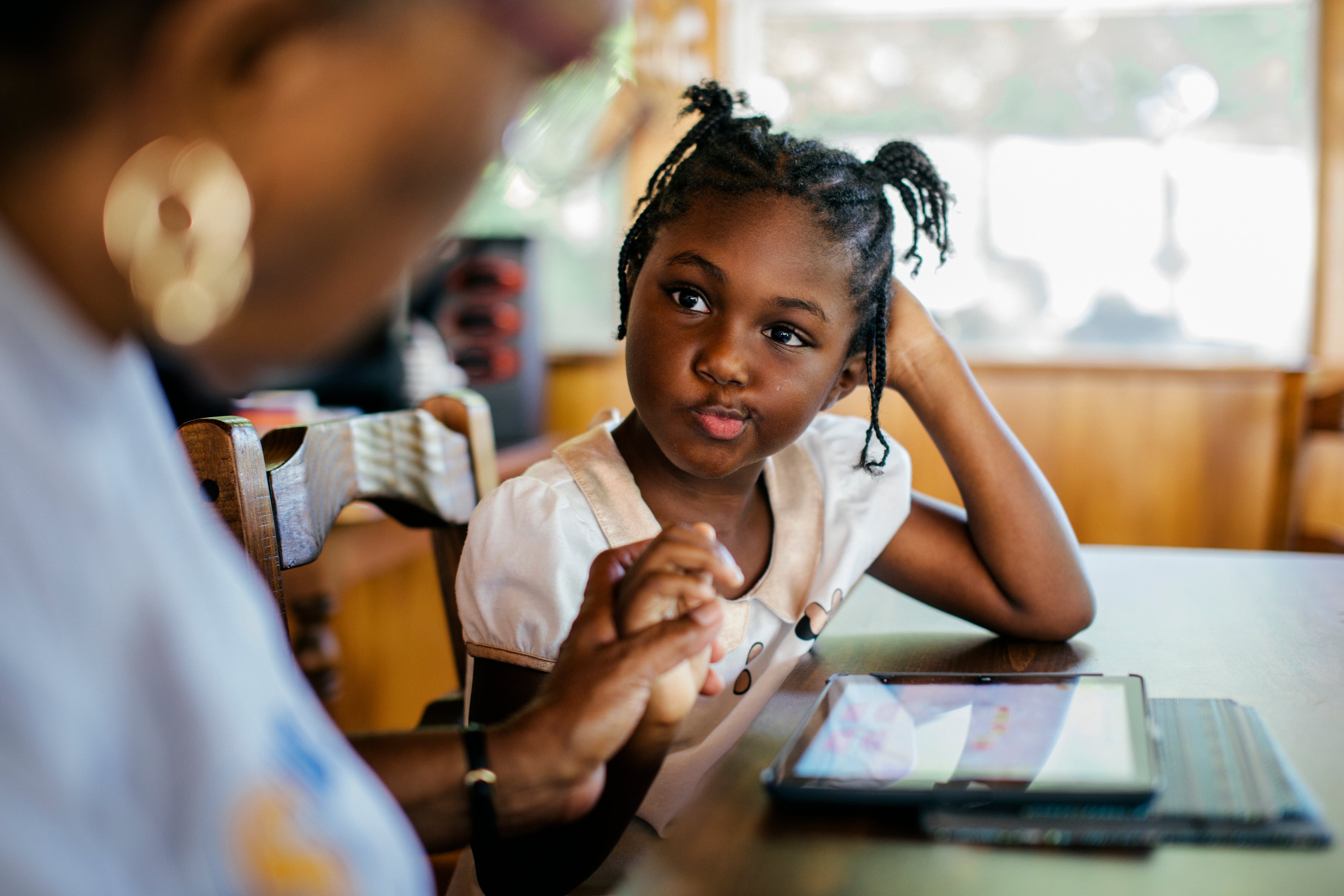 A young girl with braided hair works on a tablet with her grandmother. She has an inquisitive look on her face as she rests her head on her hand.